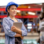 Portrait of factory worker woman with blue hardhat holds tablet and stand in spare parts workplace area. Concept of confident of working with spare parts planning software.