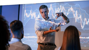 Company growth plan. Male leader talking to employees, showing information on the projector in office of stock exchange company